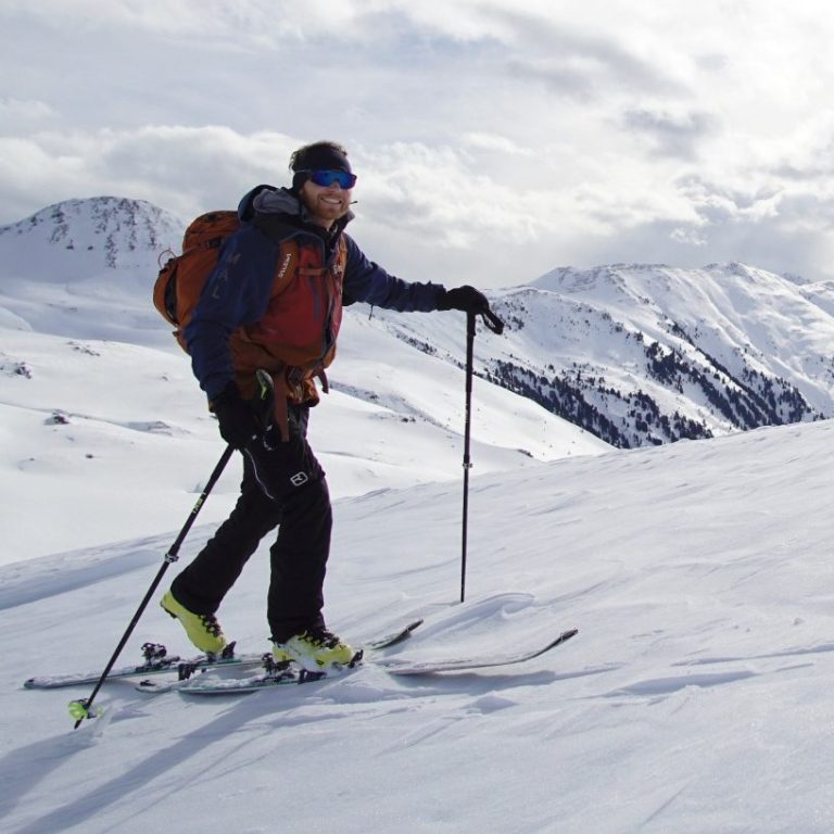 Skitour in Kitzbühel Skifahrer mit Rucksack auf verschneitem Berg, umgeben von Schnee und Wolken.