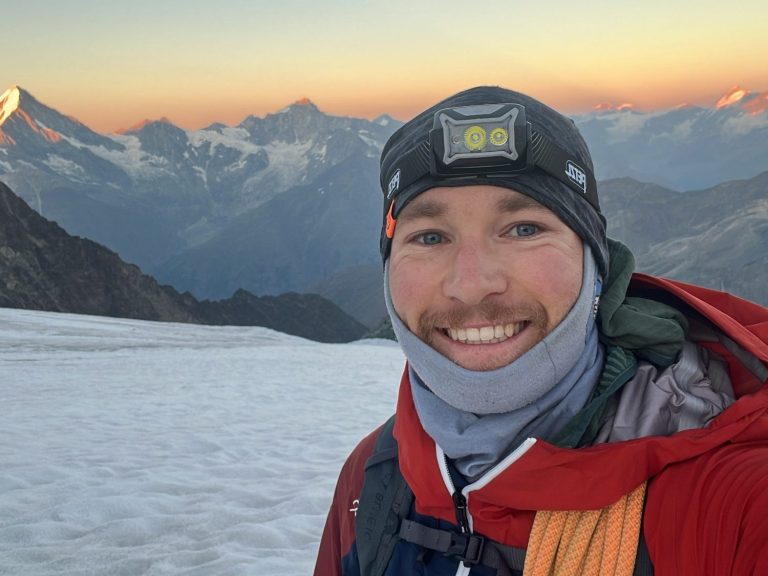 Max im Wallis Mann in warmer Kleidung, lächelt vor schneebedeckten Bergen bei Sonnenaufgang.